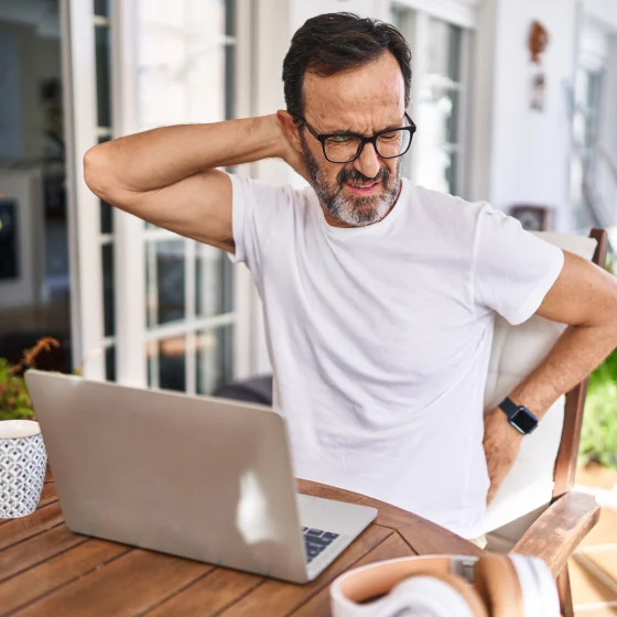Middle age man using a laptop at home suffering of neck ache. He's touching his neck with his hand and seems to have discomfort.